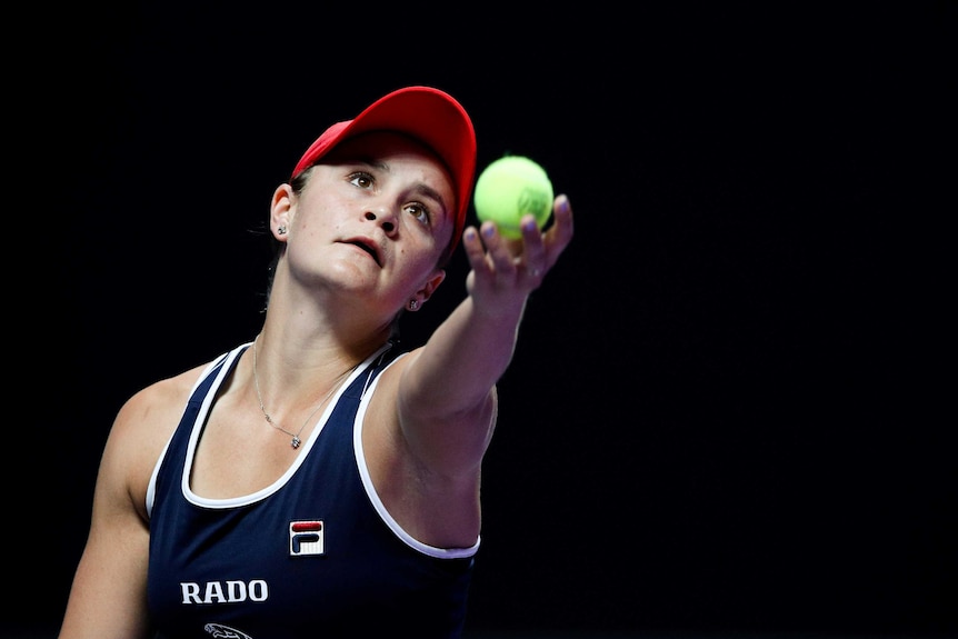 Ash Barty about to toss the ball up to serve, with a dark background.
