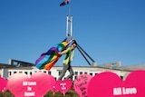 Person waves rainbow flag at Parliament House in Canberra. Heart signs saying 'All love is equal' are in the forground