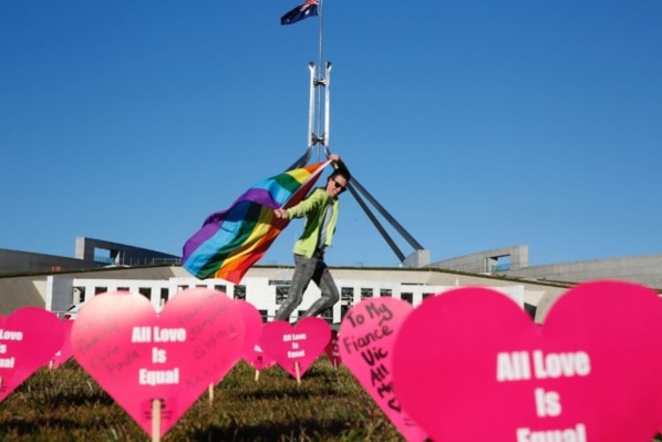 Person waves rainbow flag at Parliament House in Canberra. Heart signs saying 'All love is equal' are in the forground