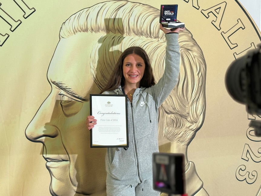 A girl holds a collectible coin in the air with a certificate saying it is the first minted in 2024.