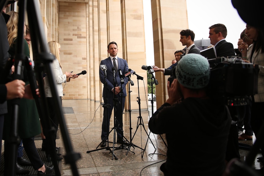 A man in a suit shot form a low angle with journalists and microphones crowded around him outside of parliament.