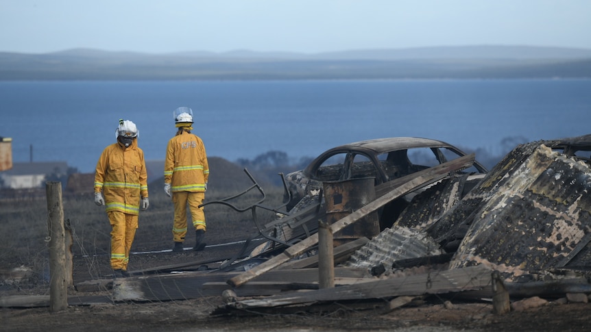 Two firefighters walk in opposite directions next to a burnt out car and rubble