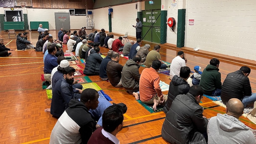 Dozens of Muslim men engage in prayer on an indoor basketball court.