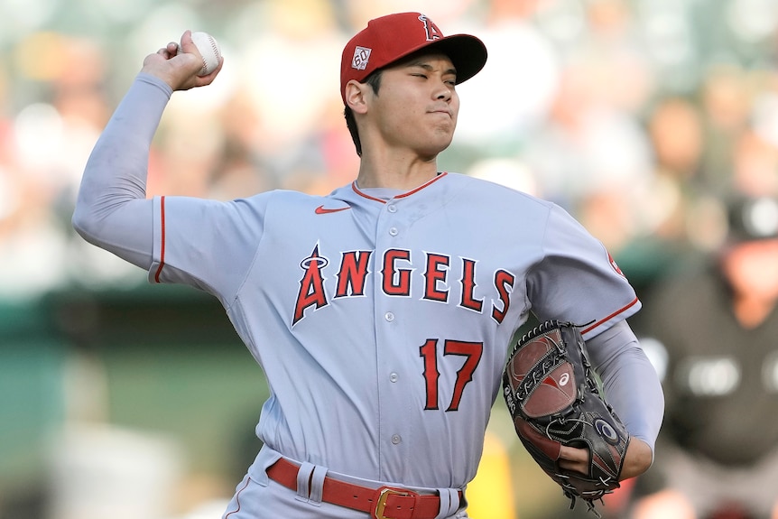 A Japanese man in a grey baseball uniform and red hat pitches a baseball with his right arm