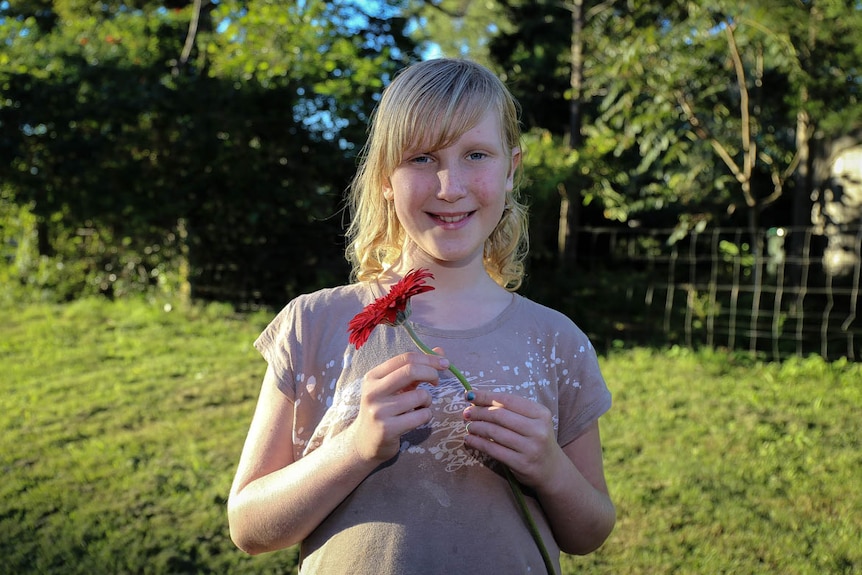 A girl with shoulder-length blonde hair, wearing a mauve t-shirt, smiling and holding a flower.