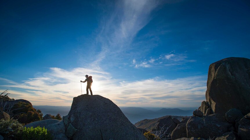 A hiker stands on a big granite rock with the sun and blue sky and white clouds behind him on Mt Buffalo.
