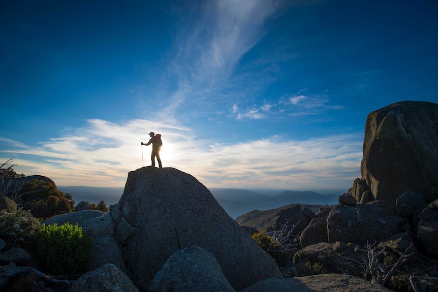 A hiker stands on a big granite rock with the sun and blue sky and white clouds behind him on Mt Buffalo.