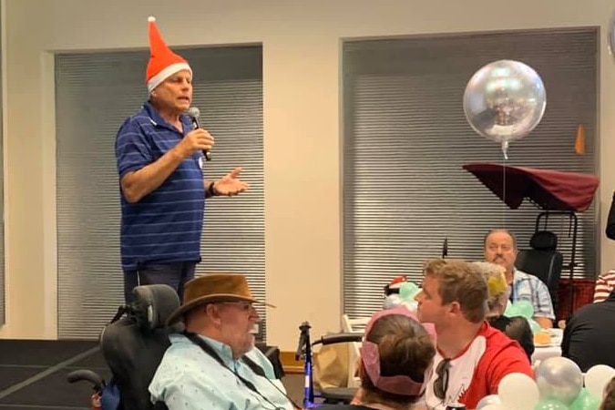 A man in a Christmas hat speaks into a microphone while people eat lunch in town hall