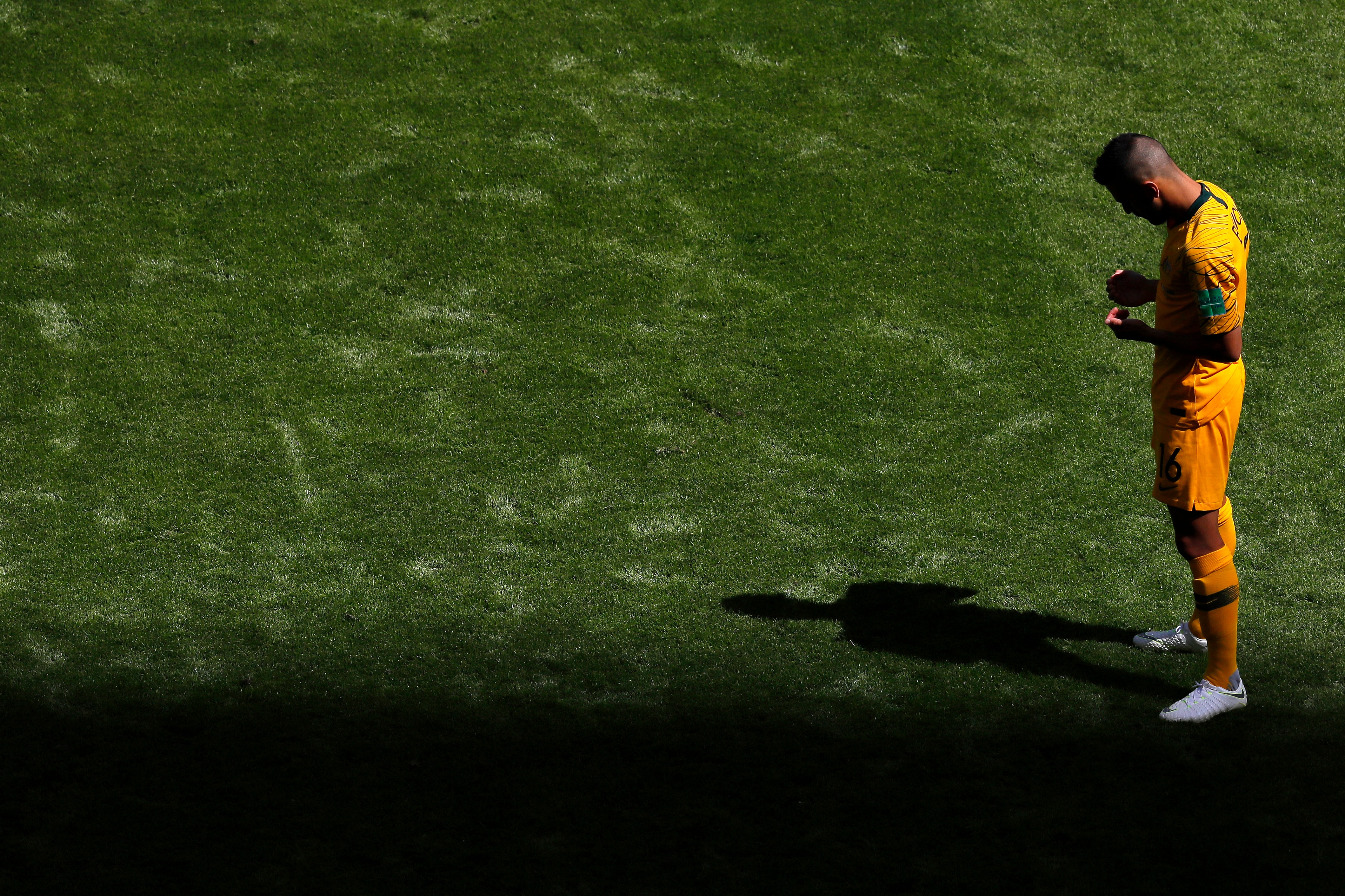 A male soccer player wearing yellow looks down at his hands while standing on grass