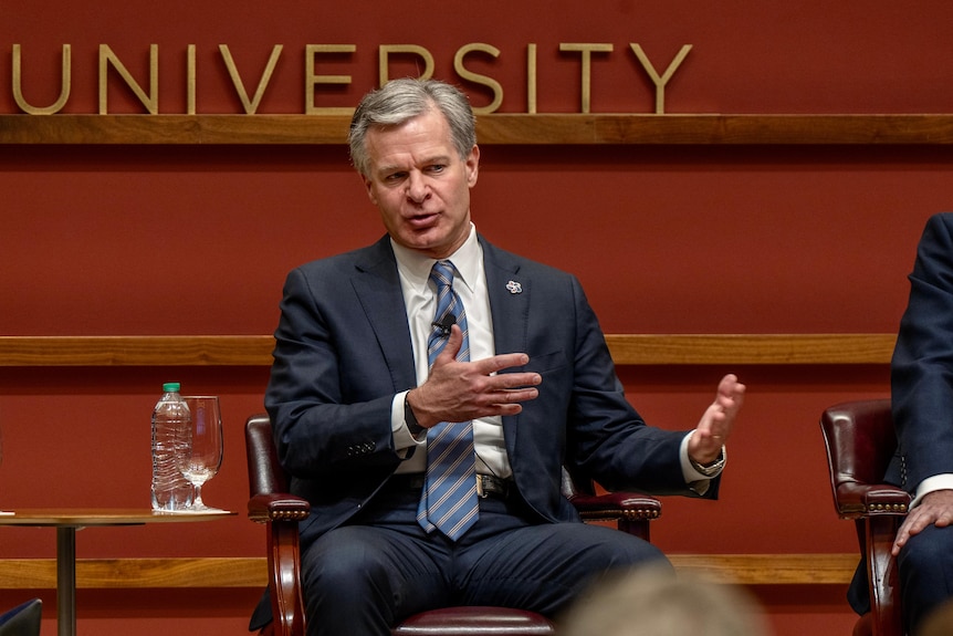 Mike Wray is seated in front of a brown all. He is speaking and gesturing with his hands.