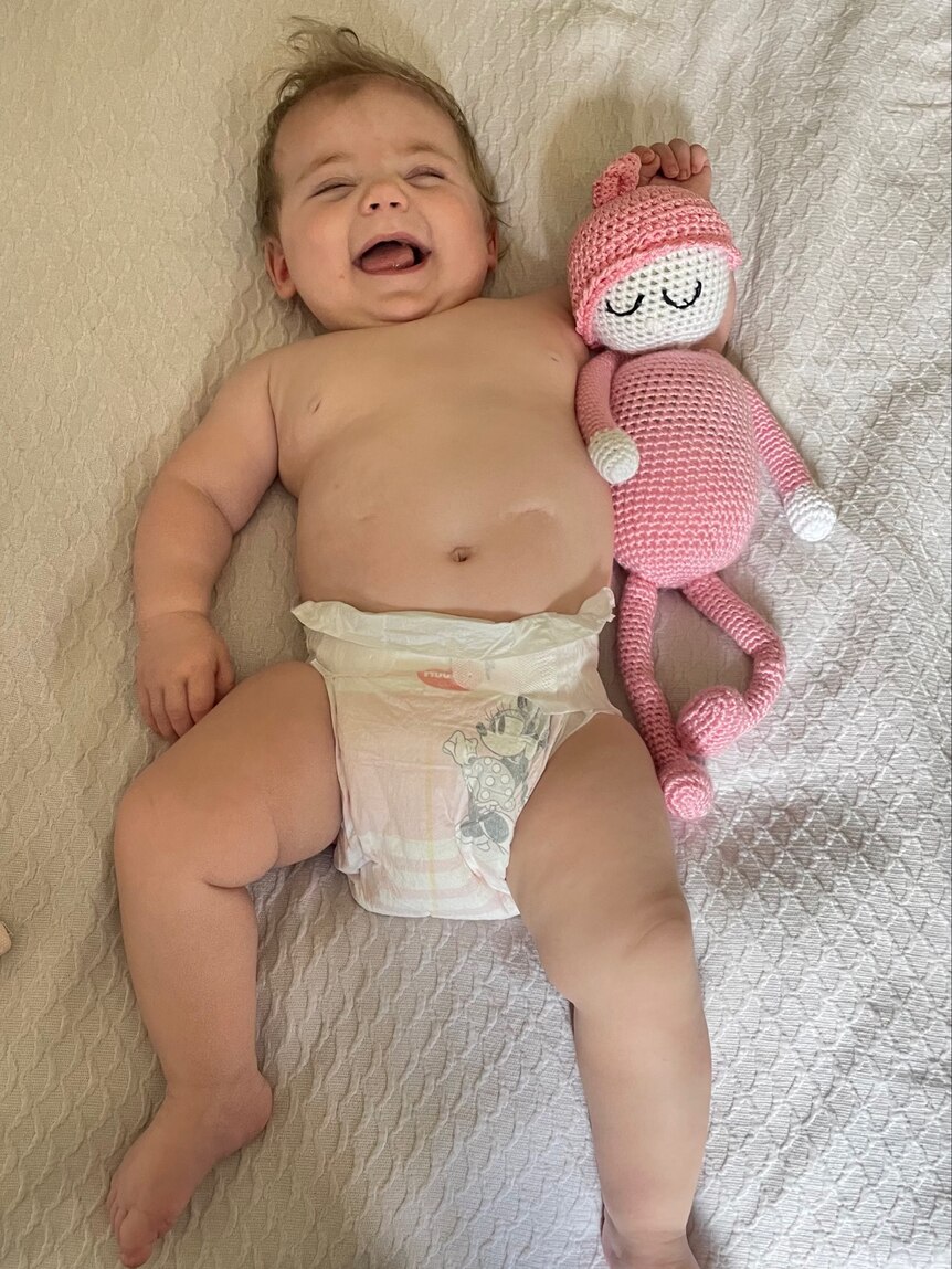 A laughing baby wearing a nappy beside a pink knitted doll, in a cot. 