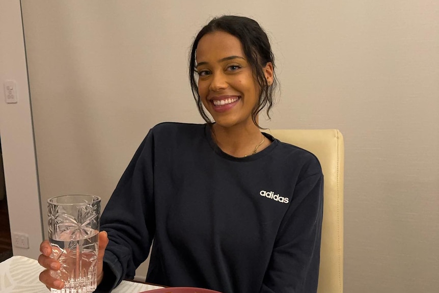 A photo of a woman wearing a black long sleeve shirt, holding a glass of water with a plate of food in front of her.