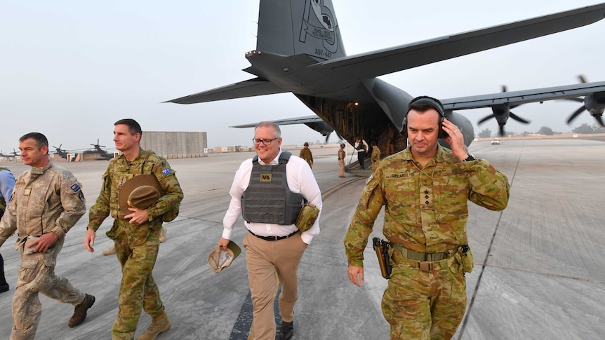 Scott Morrison, wearing a flak jacket, walks away from a Defence plane with men in camouflage uniforms.