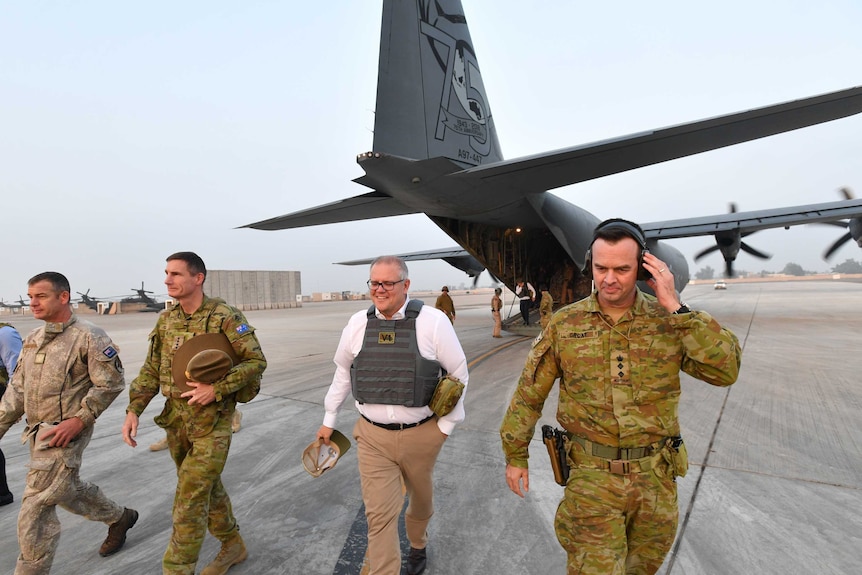 Scott Morrison, wearing a flak jacket, walks away from a Defence plane with men in camouflage uniforms.