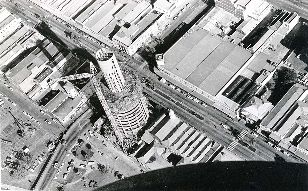 A black and white photograph taken from a helicopter captures the construction of a circular high rise building in the 1970s.