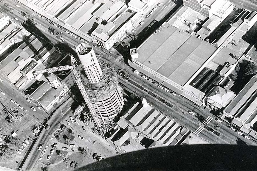 A black and white photo taken from a helicopter shows the construction of a circular high-rise building in the 1970s.