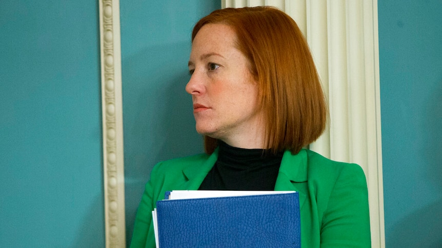 A woman with a red bob wears a green jacket and carries a blue folder and a pen.
