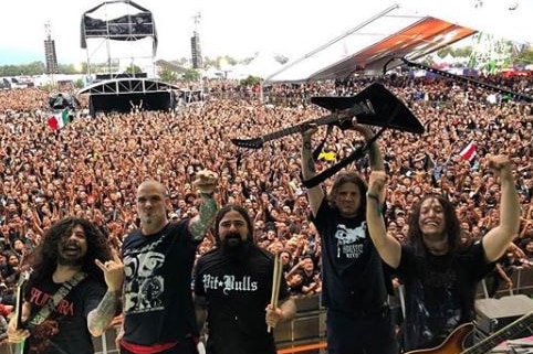 Five members of a metal band on stage with their backs to a big crowd, one with their guitar above their head