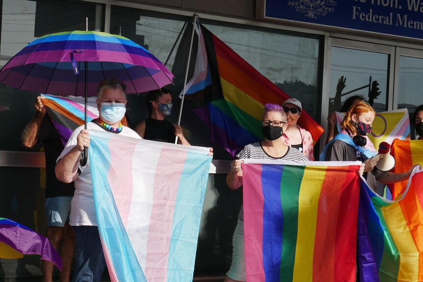 A crowd of people outside an office holding trans and LGBT flags and placards in protest.