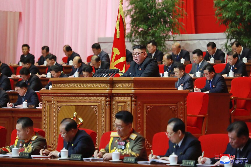 North Korean leader Kim Jong Un speaks from a podium surrounded by party members dressed in uniform and writing notes.