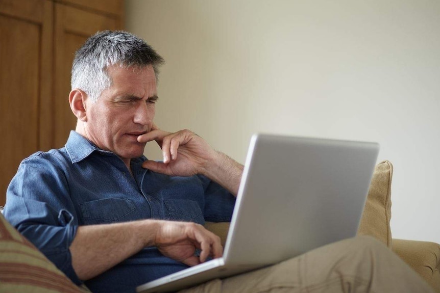 A man sits on an armchair with a laptop on his lap, and his face is screwed up as if confused.