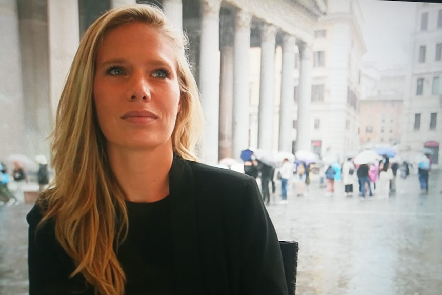 A woman with blonde hair looking off camera with people in square under umbrellas in blurry background behind her.