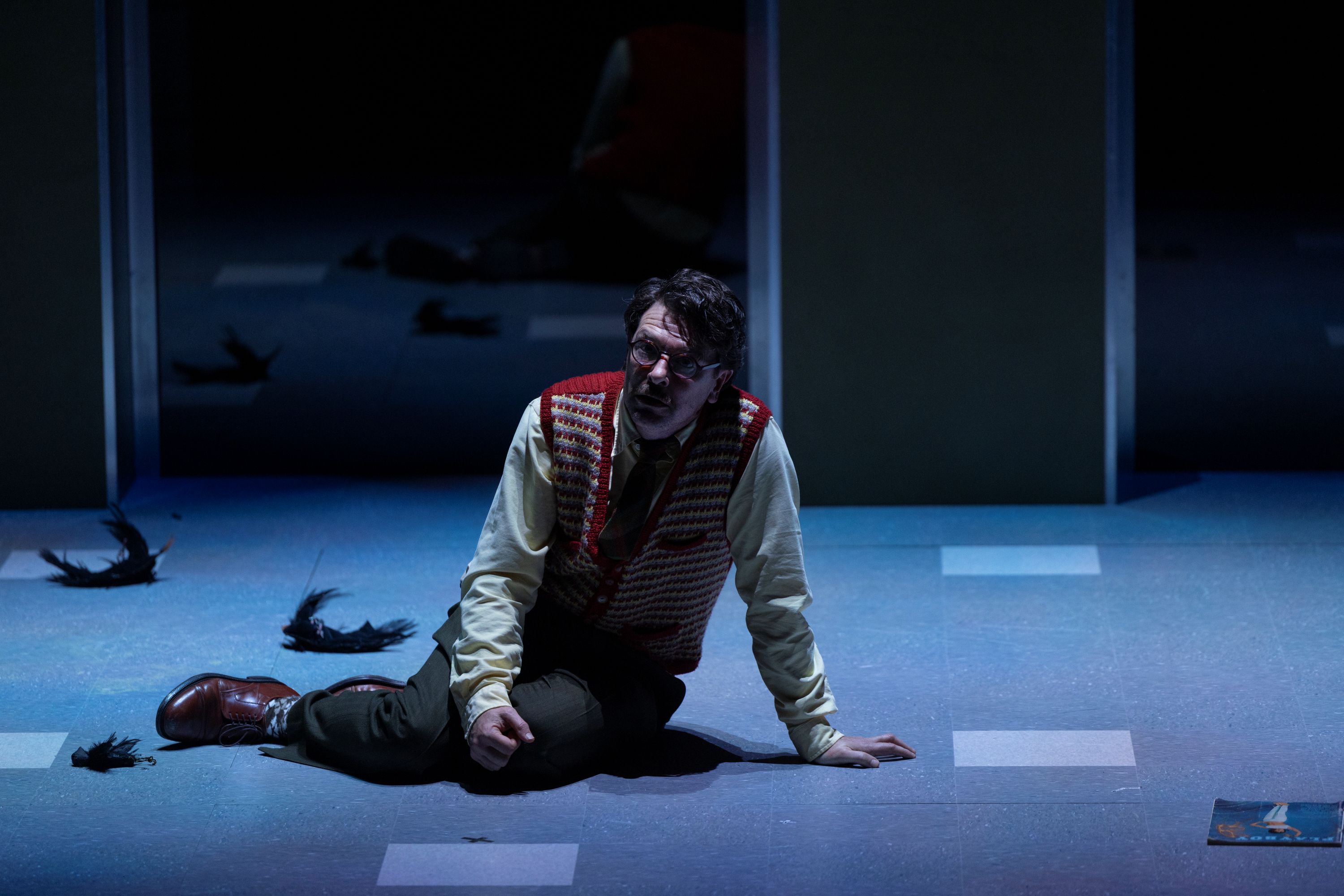 On stage, Dan Spielman, 47, in glasses, is sitting on his side on the floor, under dim lighting, looking distressed.