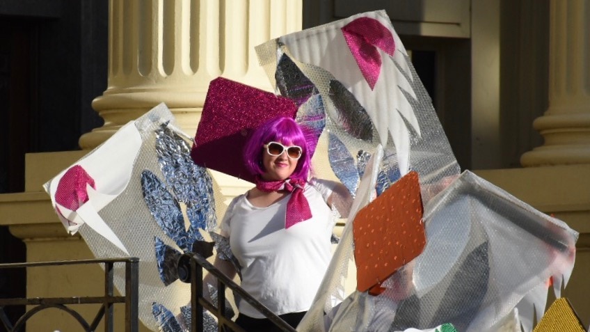 A woman with a purple wig and sunglasses holds colourful banners of patched material.