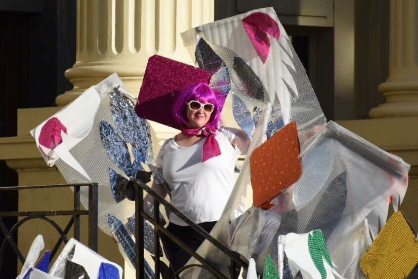 A woman with a purple wig and sunglasses holds colourful banners of patched material.