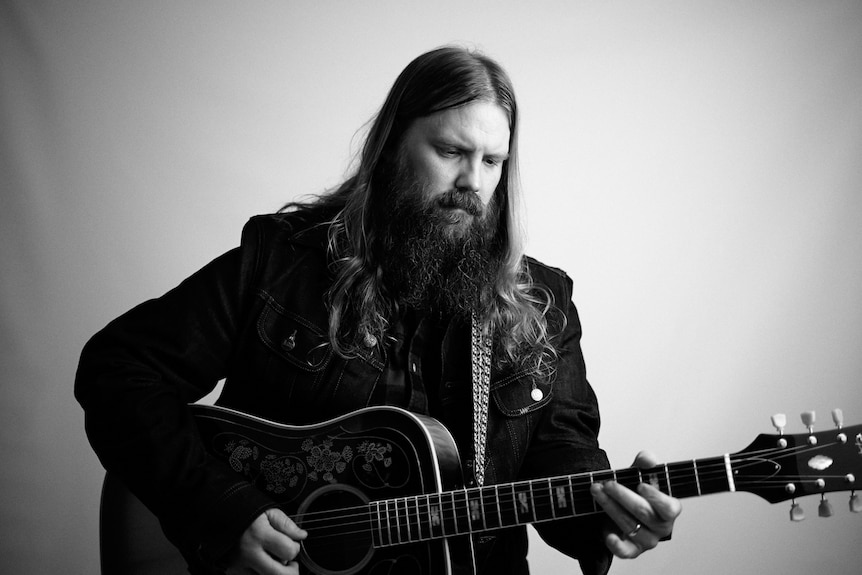 A black and white photo of a man with long hair and beard playing a guitar. He is wearing a jacket. 