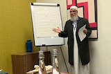 A man in Islamic wear speaking to a room. 