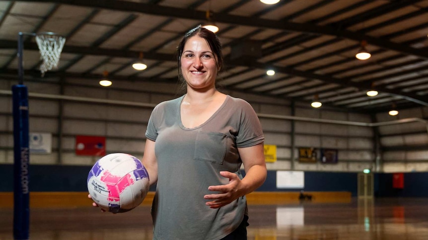 Kelly is wearing a grey T-shirt and holding a netball ball while standing on a netball court.