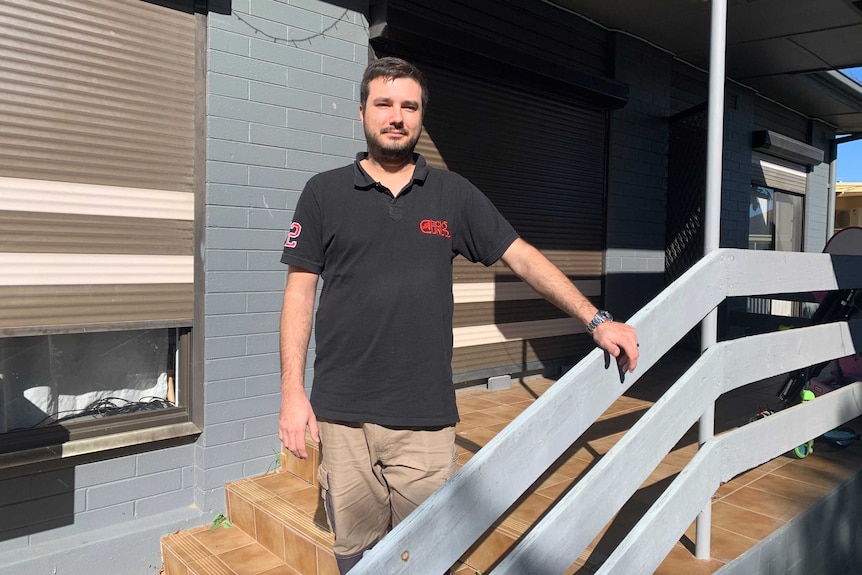 Chris Pascoe, wearing black polo shirt and long brown shorts, standing on the front steps of a grey brick house