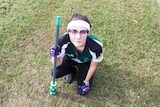 Young woman crouched down on grass holding 'broomstick', gloves and wearing quidditch outfit.