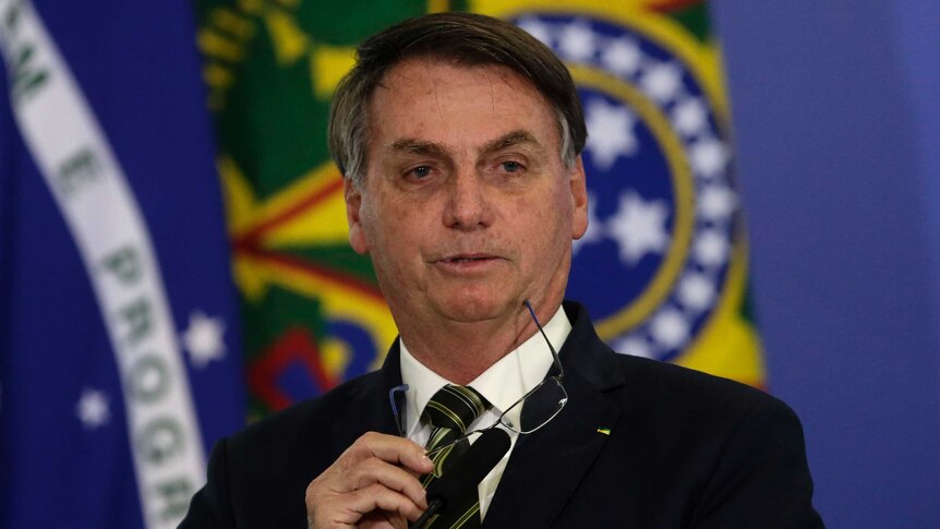 Brazil's President Jair Bolsonaro takes off his glasses as he speaks in front of a microphone with flags in the background.