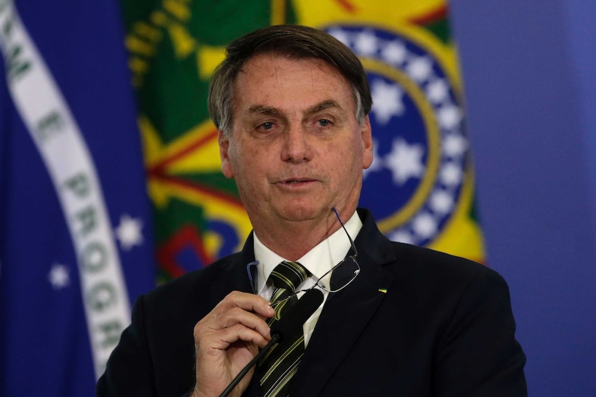 Brazil's President Jair Bolsonaro takes off his glasses as he speaks in front of a microphone with flags in the background.