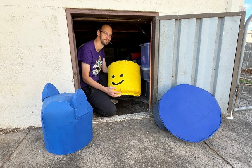 A man squatting in the doorway of a basement storage area with three large Lego heads, wears glasses, has beard.