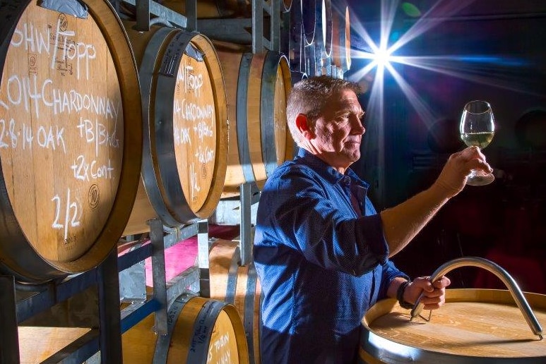 A man stands in a room with barrels holding a glass of wine.
