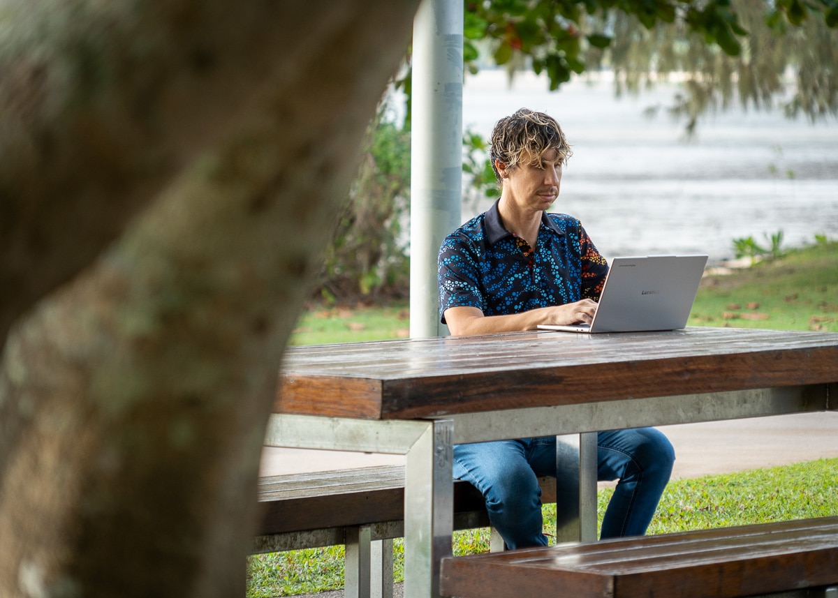 A young adult male looking at his laptop on a park bench