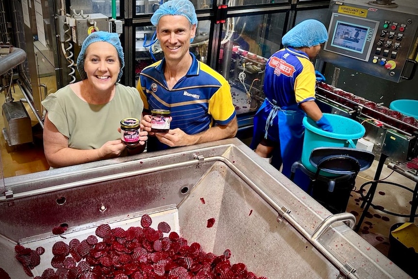 Two people smile above a vat of sliced beetroot holding cans of beetroot.