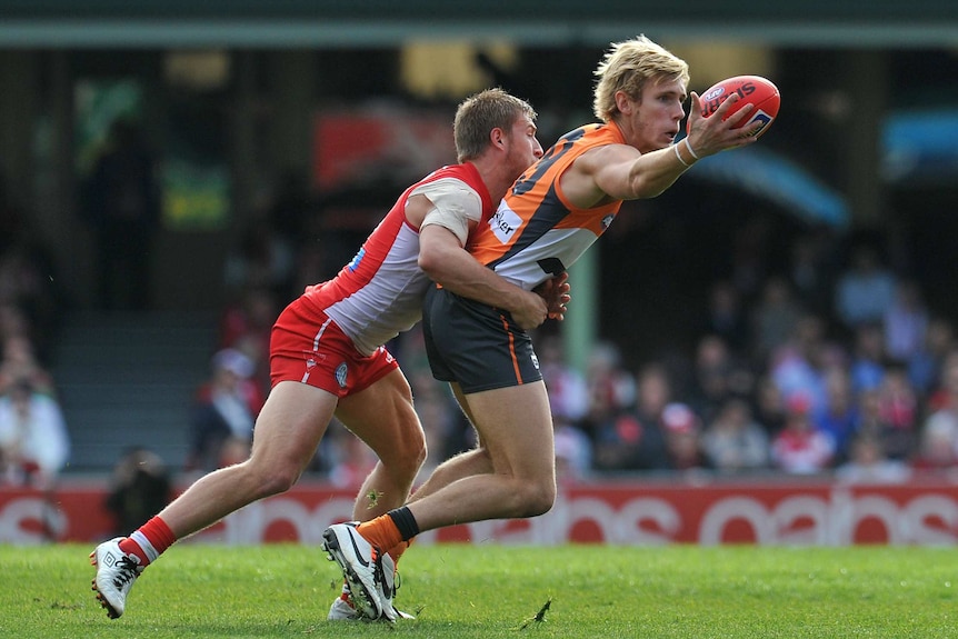 An AFL player grasps the ball in one hand as he runs forward, while an opponent tries to tackle him.