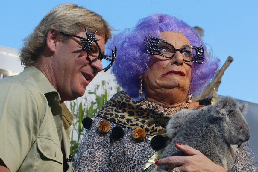 Dame Edna and Steve Irwin both in glasses stand together as Edna holds a koala and makes a face.