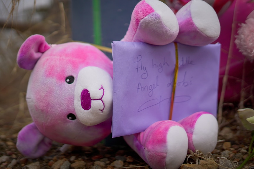 A teddy holding an envelope with 'fly high little angel Kobi' written on it