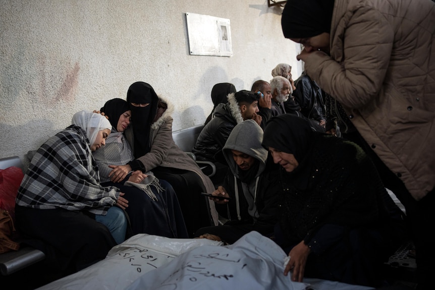 A group of mourning women sit on a bench as others examine shrouded bodies. 