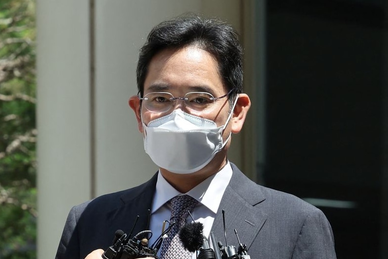 A man with short black hair wearing a suit, face mask and clear glasses being questioned by media outside on a bright day