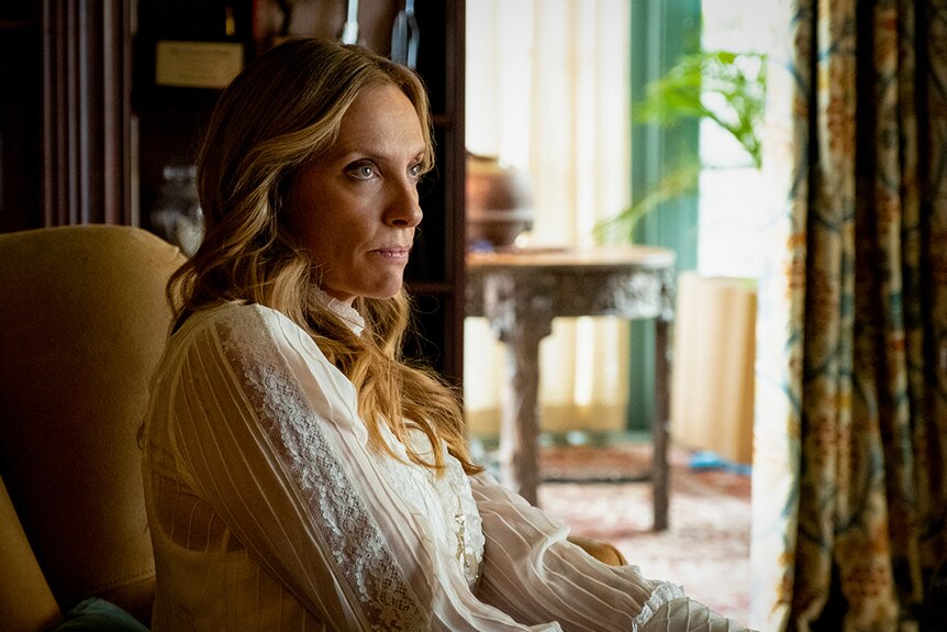 Toni Collette in white silk blouse sits on mustard armchair in dark wood and ornate interior on a sunny day.