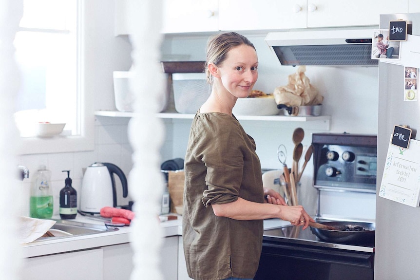 A woman stands in her home kitchen, looking at the camera, and stirring a fry pan full of mushrooms.