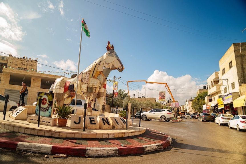 A roundabout in the middle of a road includes a sign for JENIN and a sculpture of a horse
