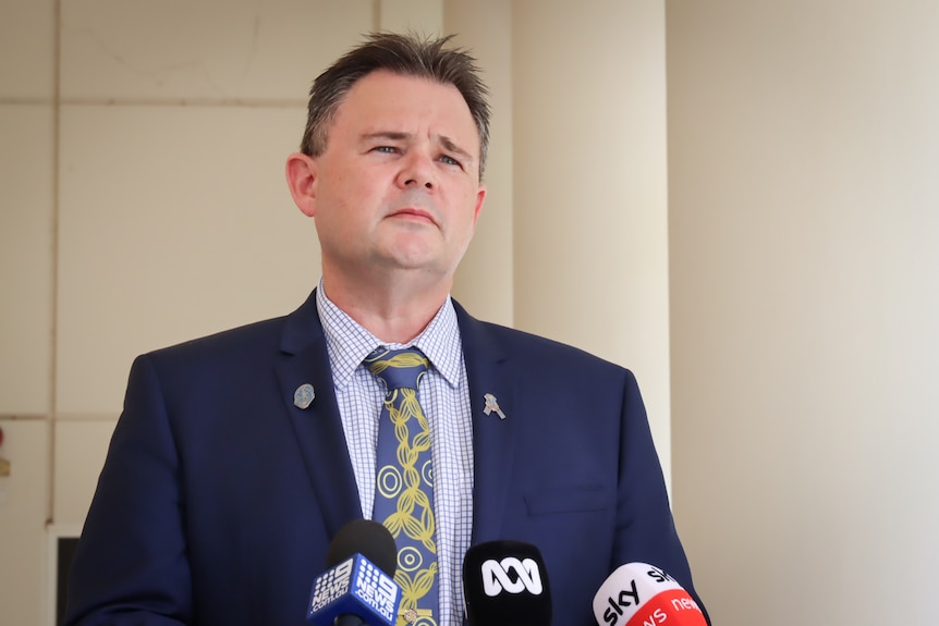 A man wearing a navy suit stands in from of microphones. He looks serious.