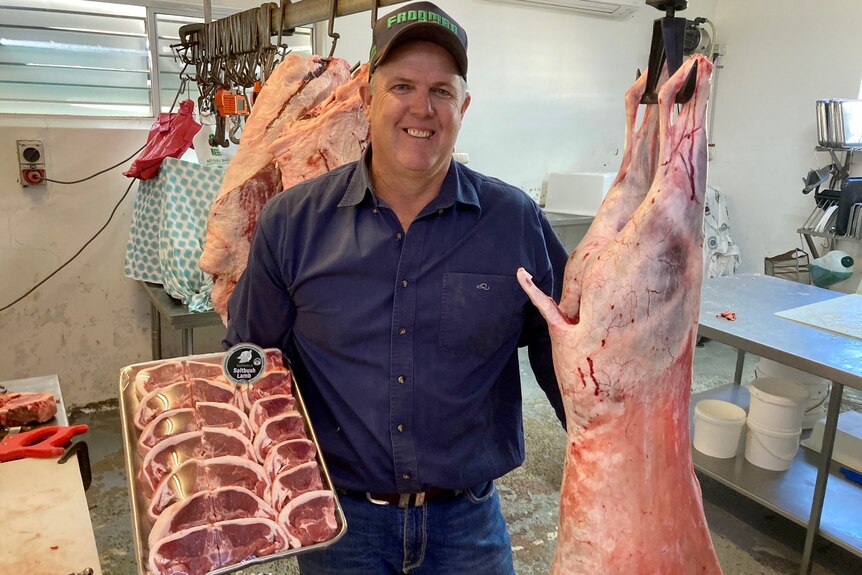 A man in a blue shirt and cap smiles while holding a tray of meat and a carcass in a butcher shop.